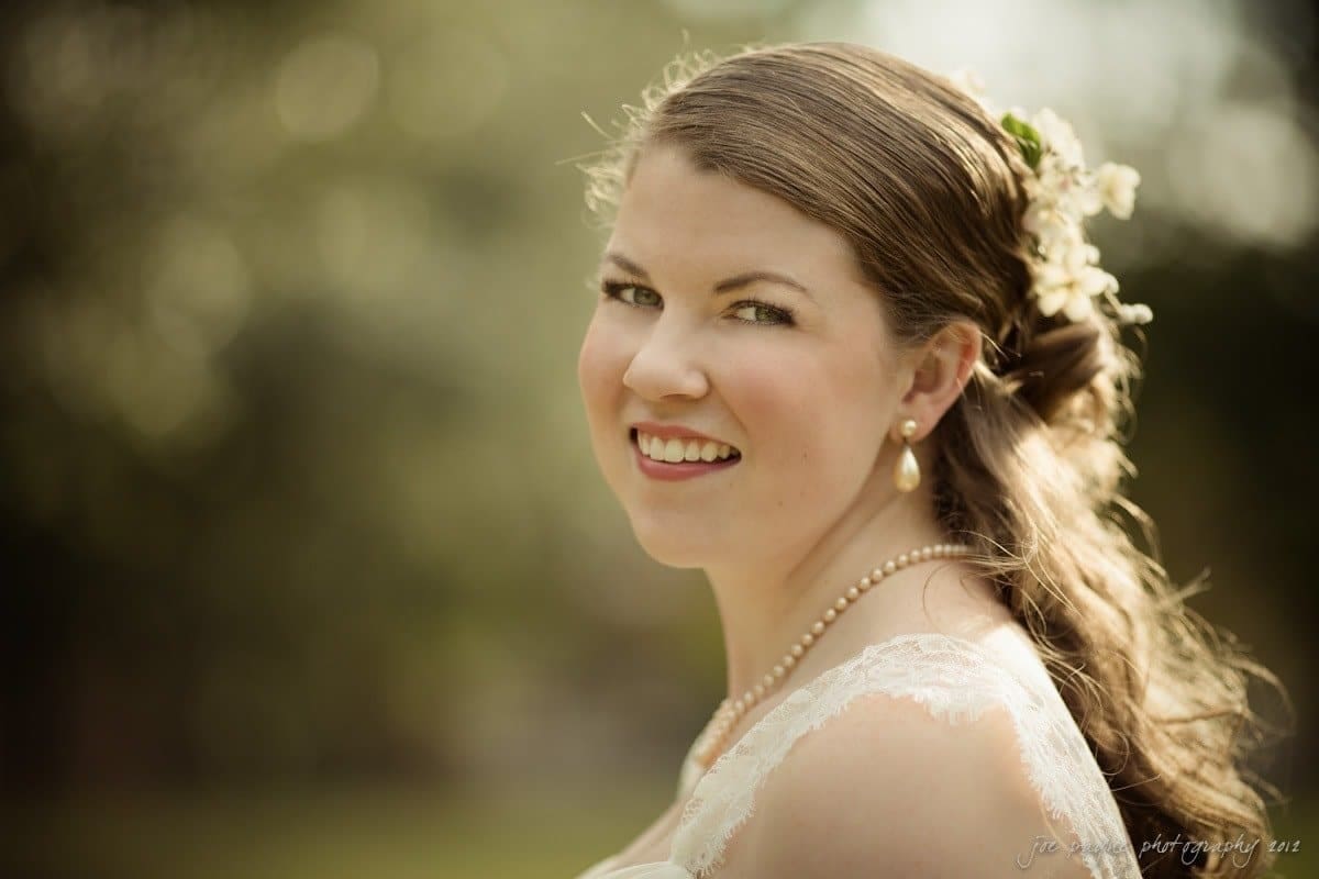A smiling woman with long hair adorned with white flowers, wearing pearl earrings and a necklace, and a lacy top. She is outdoors, with a blurred, natural background.