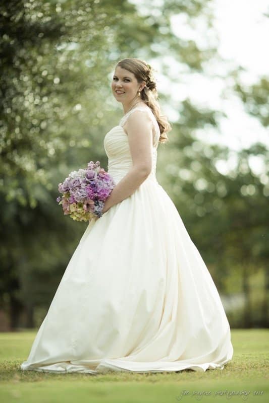 A bride in a white wedding dress stands outdoors, holding a bouquet of purple and pink flowers. She smiles softly, with trees and greenery in the blurred background. The setting is sunlit.
