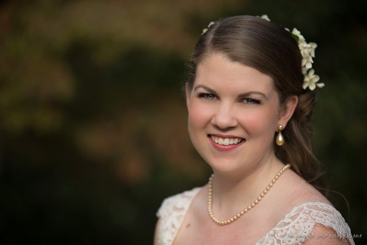 A bride with a pearl necklace and floral hair decoration smiles in an outdoor setting, wearing a lace dress. The background is softly blurred, highlighting her joyful expression and elegant attire.
