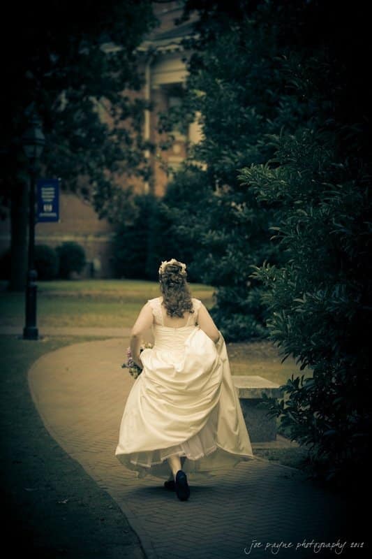 A bride in a white dress walks briskly down a tree-lined brick path, holding her gown. She has curly hair and a flower crown. The setting is outdoors with a blurred building in the background.