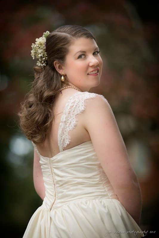 A bride with long, wavy hair adorned with flowers poses outdoors in a white wedding dress with lace straps. She looks over her shoulder with a soft smile. The background is blurred with hints of greenery and red foliage.