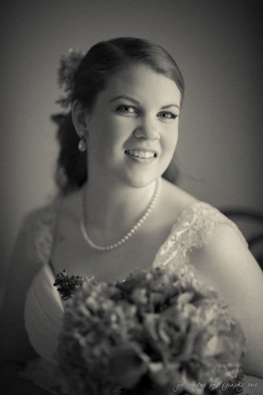 A woman in bridal attire smiles softly, holding a bouquet. She wears a lace dress with a pearl necklace, and her hair is adorned with flowers. The black and white image has a soft, classic ambiance.