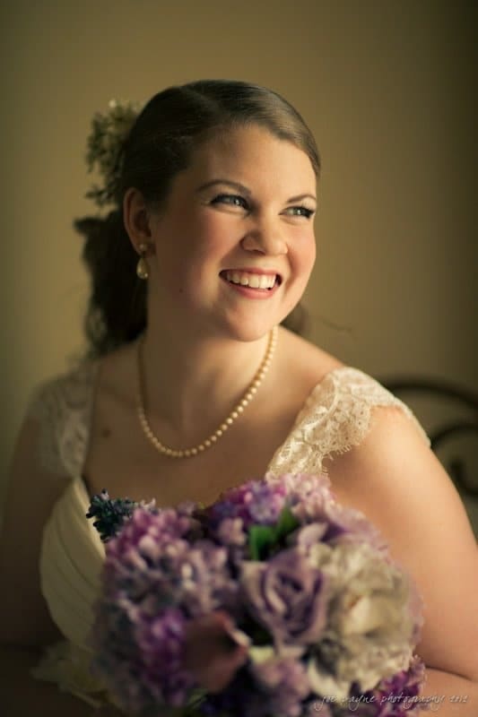 A smiling bride holds a bouquet of purple and white flowers. She is wearing a lace wedding dress and a pearl necklace, with her hair styled back. Soft, natural light illuminates her face, creating a warm, joyful atmosphere.