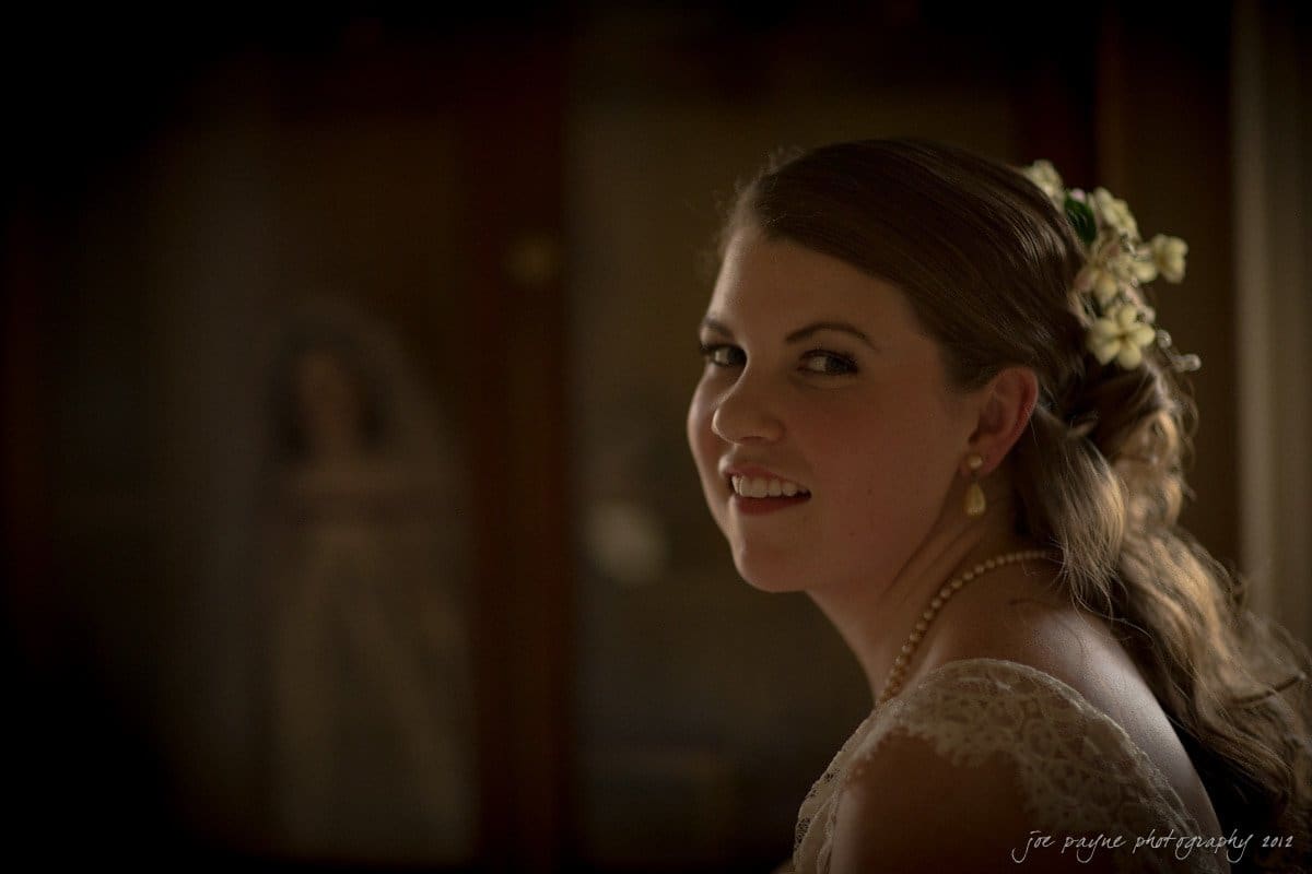 A bride with a serene smile looks over her shoulder. She has delicate flowers in her hair and wears lace-trimmed attire with a pearl necklace. The background is softly lit, creating a warm, intimate atmosphere.
