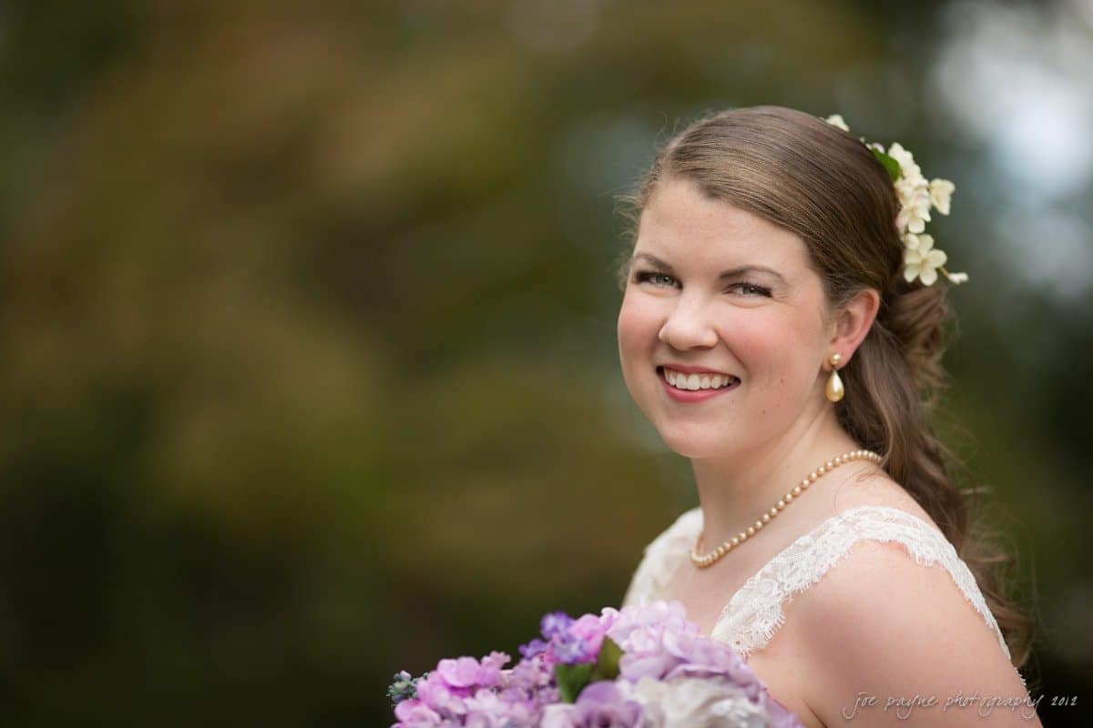 A bride with long brown hair adorned with flowers smiles broadly outdoors. She holds a bouquet of purple and white flowers and wears a pearl necklace with a lace wedding dress. The background is a blurred natural setting.