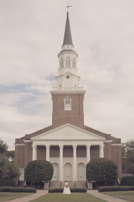 A brick church with a tall white steeple and a clock faces the viewer. The entrance features four columns and a triangular pediment. A person in a white outfit stands on the path in front, surrounded by neatly trimmed bushes. The sky is cloudy.