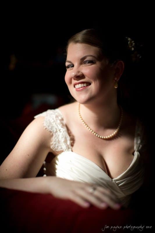 A woman in a white dress smiles warmly while sitting in a softly lit room. She wears a pearl necklace and pearl earrings, and her hair is styled elegantly. She rests her arm on a nearby surface, creating a relaxed and joyful atmosphere.