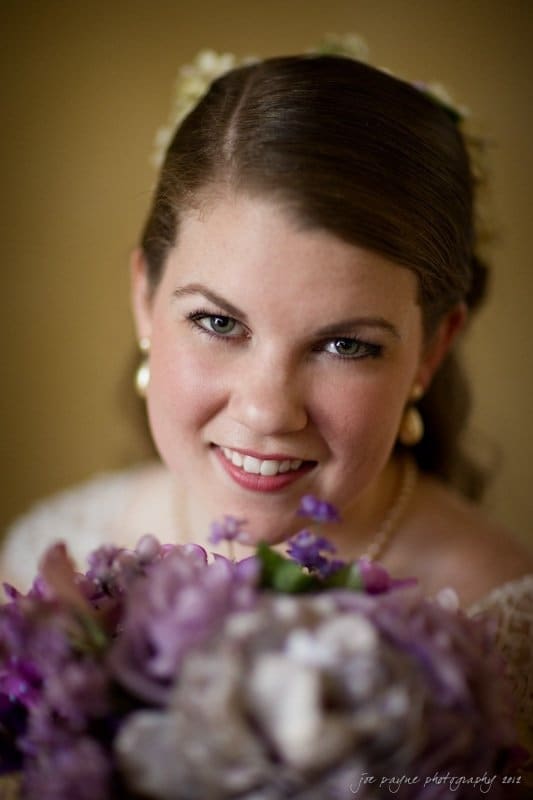 A bride with a gentle smile holds a bouquet of purple flowers. She wears pearl earrings and a white dress, with her hair styled elegantly. The background is softly blurred, emphasizing her joyful expression.