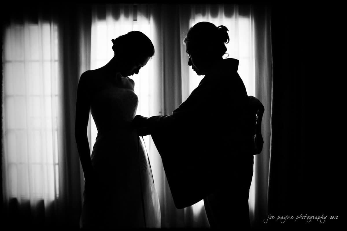 A bride in a wedding dress stands gracefully as another person, possibly her mother, adjusts her gown. They are silhouetted against a large window, with soft light creating an intimate and serene atmosphere.