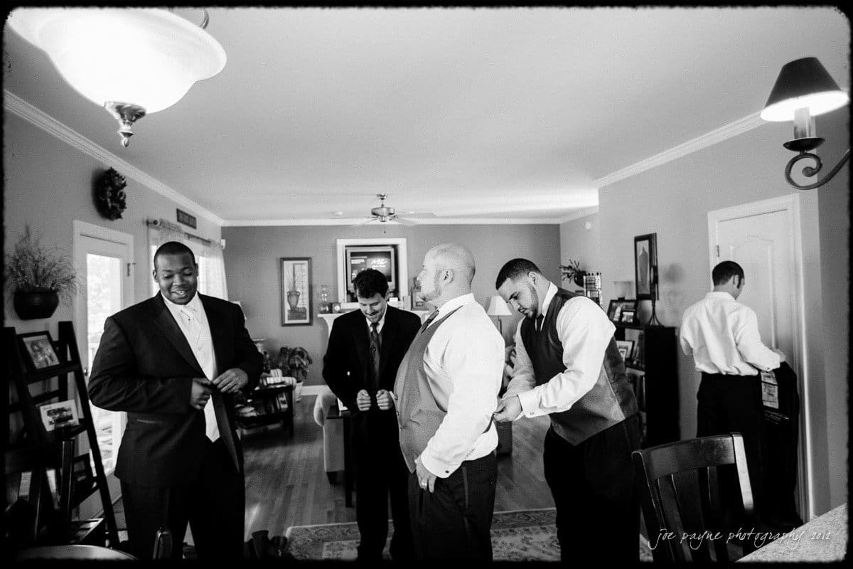 A black-and-white photo of a group of five men dressed in formal suits, preparing for a wedding in a living room. One man helps another adjust his vest, while others button their jackets. The room is decorated with framed photos and plants.