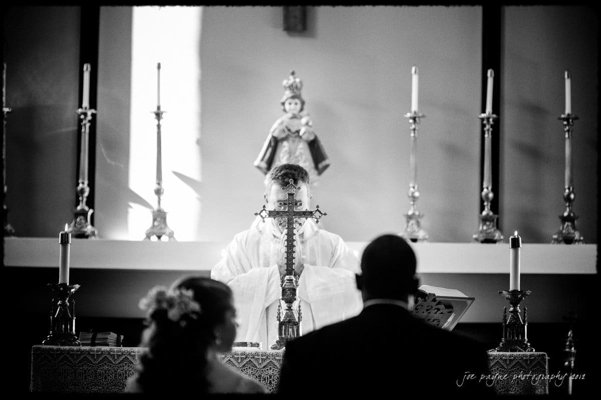 A priest in white robes conducts a religious ceremony at an altar in a church. The altar is adorned with tall candlesticks and a crucifix. Two people stand in the foreground, facing the priest. The image is in black and white.