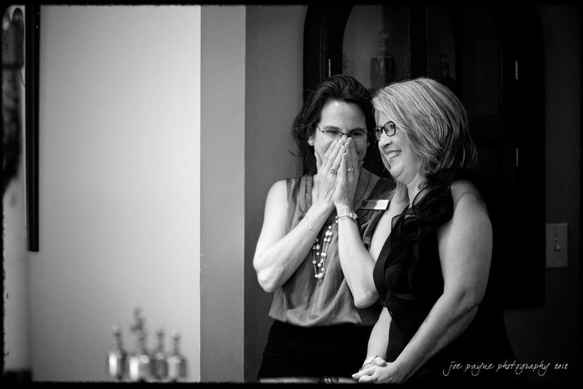 Two women stand indoors, smiling and covering their mouths as if surprised or amused. One woman has her hands over her mouth, while the other stands beside her, both wearing glasses. The photo is in black and white.