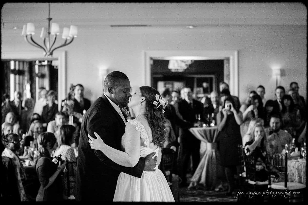 A bride and groom share their first dance in front of an audience at a wedding reception. Onlookers watch and capture the moment with phones and cameras in a warmly lit room.