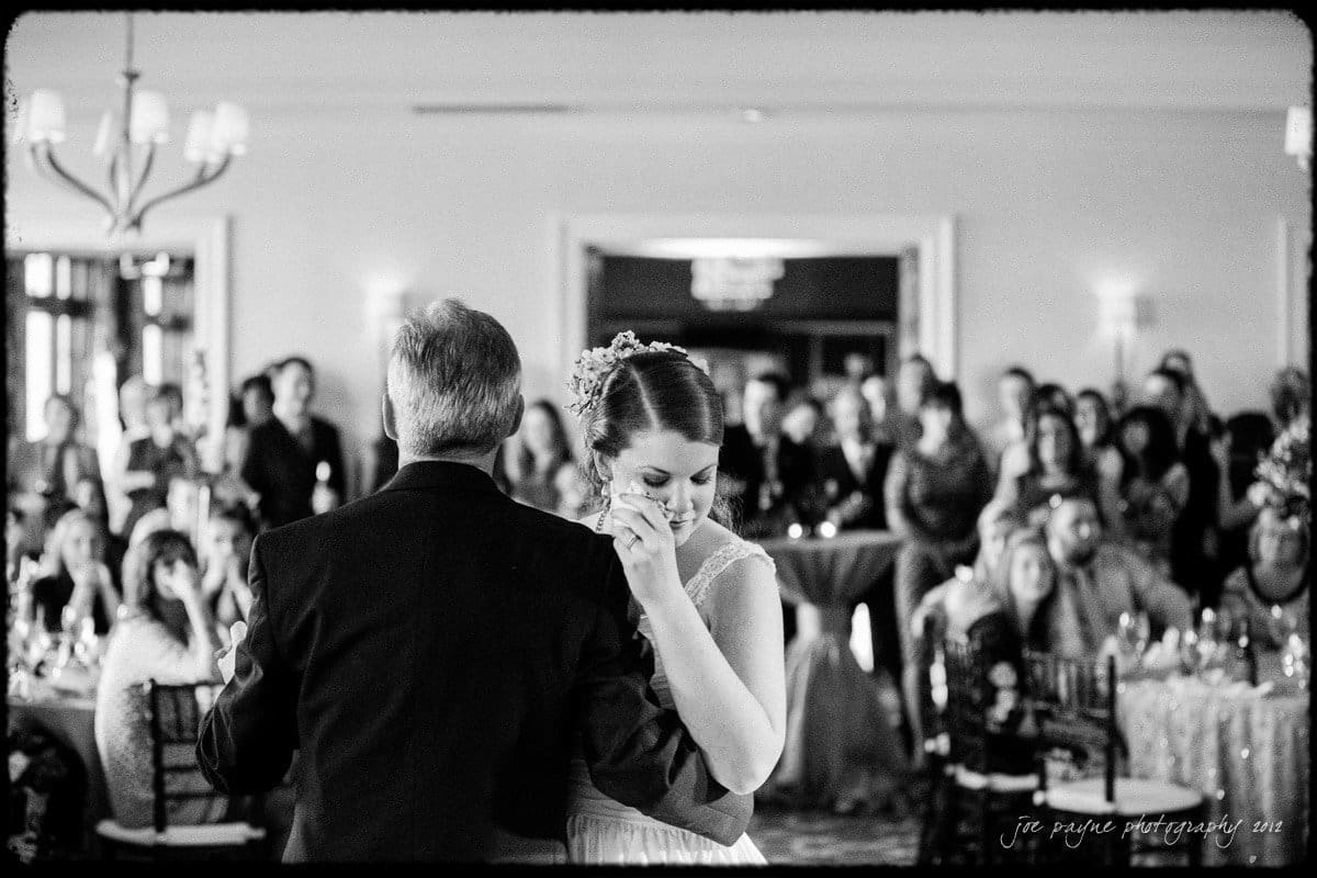 A bride dances with an older man in a warmly lit room filled with seated and standing guests. The bride wears a floral hairpiece, and the guests watch the dance attentively. The image is in black and white.