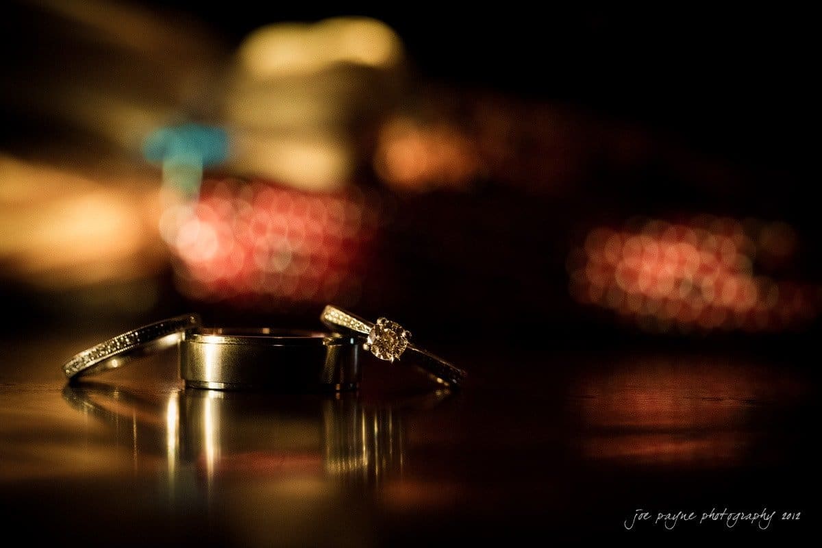 Close-up of three rings on a dark reflective surface. One is a diamond engagement ring, and the others are simple bands. The background is softly blurred with warm lighting, creating a romantic ambiance.