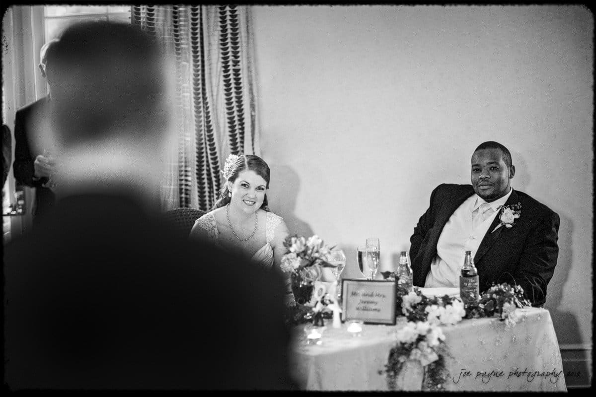 A bride and groom sit at a decorated table, smiling as they listen to a speech. The bride is wearing a white dress, and the groom is in a dark suit. The scene is captured in black and white.