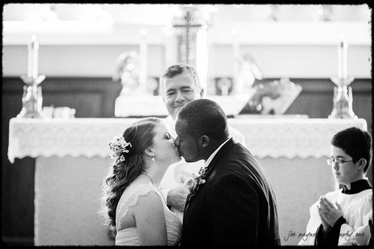 A bride and groom share a kiss at the altar during their wedding ceremony, with a smiling officiant behind them and an altar server nearby—captured beautifully by a Wake Forest wedding photographer.