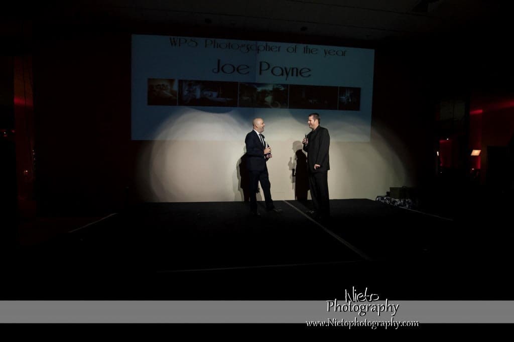 Two people stand on a dimly lit stage, one holding a microphone. A large screen behind them reads WPS Photographer of the Year Joe Payne.