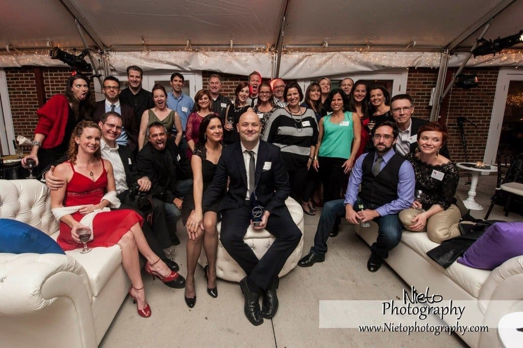 A group of people, dressed in formal and semi-formal attire, pose together at an indoor event. They are seated and standing around a man holding a trophy. The setting includes couches and a tented backdrop.