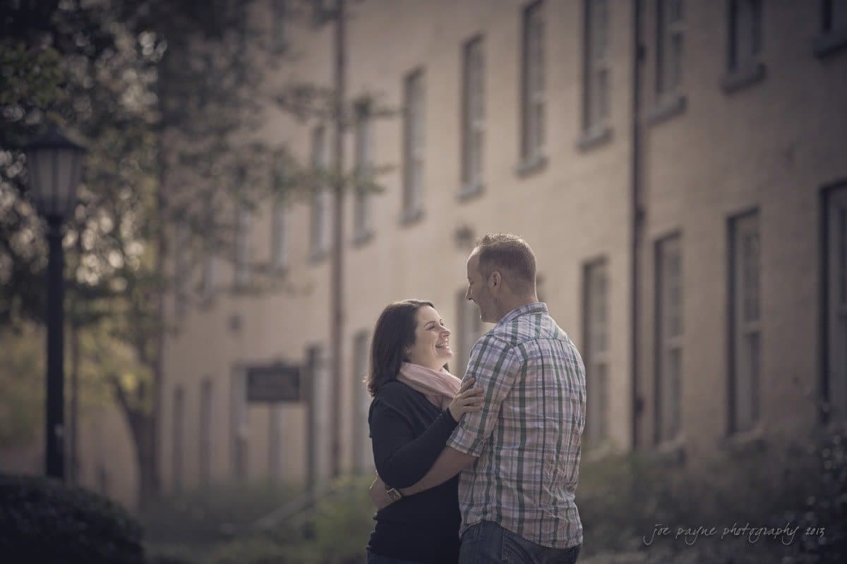 wedding photographers in raleigh nc ~ engagement session for jessica and bo A couple stands close together outdoors, both smiling at each other. They are surrounded by trees and a row of buildings in the background, creating a serene and intimate atmosphere.