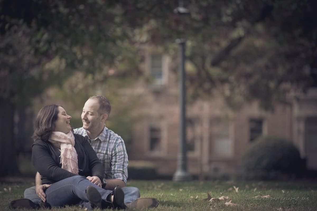 wedding photographers in raleigh nc ~ engagement session for jessica and bo A couple sits on the grass, smiling at each other affectionately. The man has his arm around the woman. They are surrounded by trees and blurred greenery in the background, suggesting a park or garden setting.