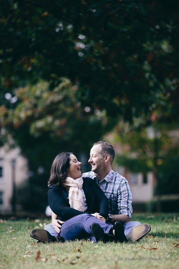 wedding photographers in raleigh nc ~ engagement session for jessica and bo A couple sits on a grassy field, smiling at each other. The woman wears a scarf, and the man is in a plaid shirt. Trees with green leaves are in the background, with blurry buildings visible. The scene conveys a relaxed, joyful moment outdoors.