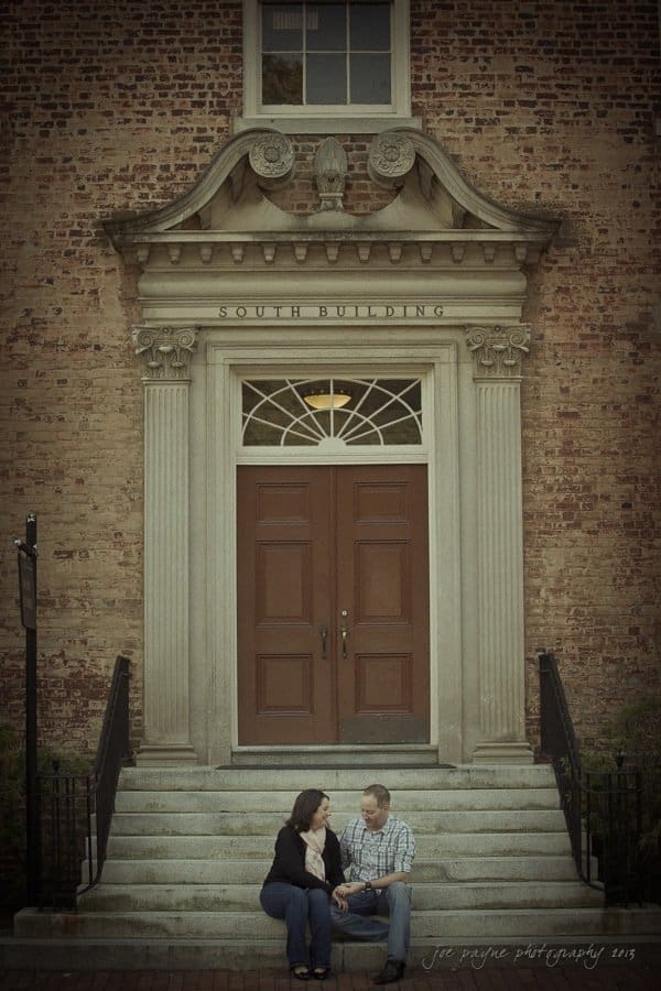 wedding photographers in raleigh nc ~ engagement session for jessica and bo A couple sits on the steps in front of a historic brick building with the words South Building inscribed above a large wooden door. They are holding hands and smiling at each other.
