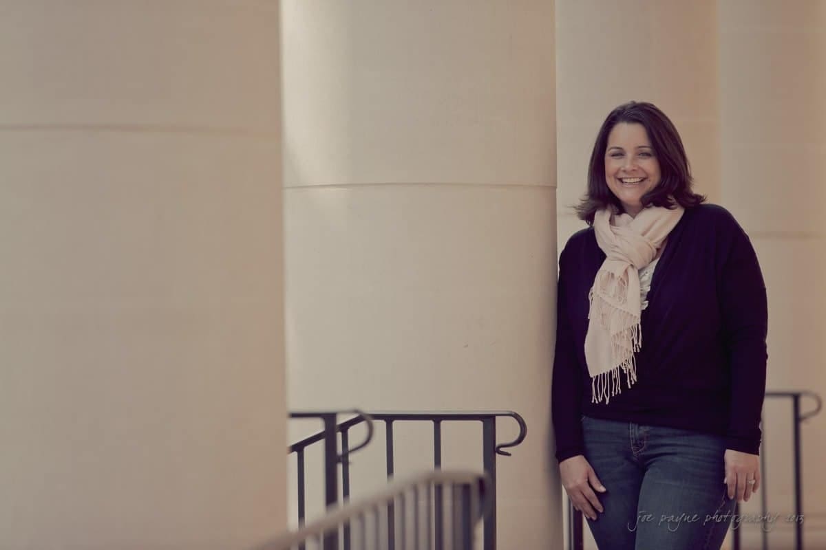 wedding photographers in raleigh nc ~ engagement session for jessica and bo A woman in a dark sweater and scarf leans against a railing between large beige columns. She smiles at the camera, with soft natural light illuminating the scene.