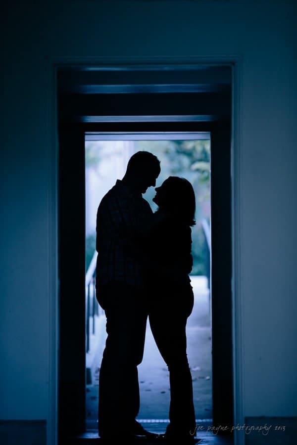 wedding photographers in raleigh nc ~ engagement session for jessica and bo Silhouette of a couple standing closely in a doorway, softly lit from behind. The scene is intimate and atmospheric, with a blurred outdoor view in the background.