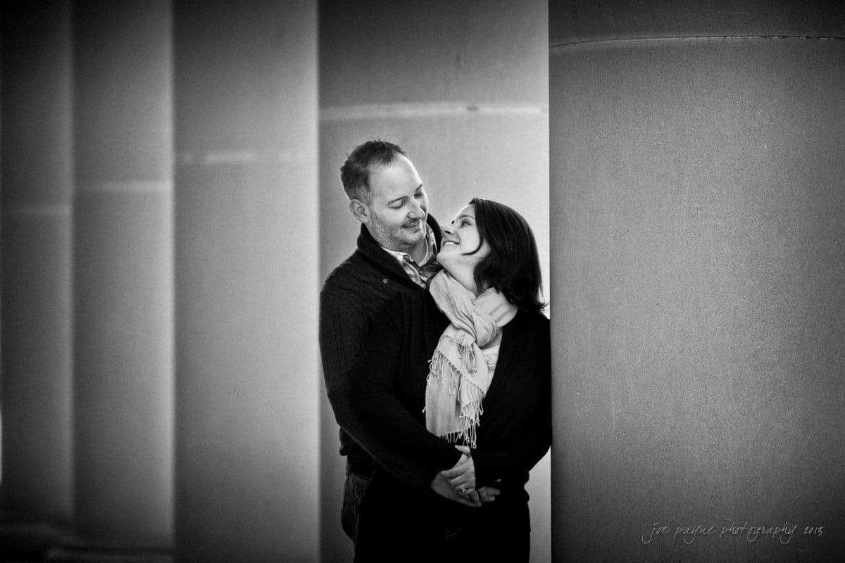 wedding photographers in raleigh nc ~ engagement session for jessica and bo A black and white photo of a couple embracing, standing between large columns. They are smiling warmly at each other. The woman is wearing a scarf, and the man has his arms wrapped around her.