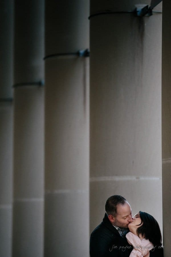 wedding photographers in raleigh nc ~ engagement session for jessica and bo A couple embraces and kisses against a tall, columned wall. The background features a series of towering vertical structures, adding depth and urban ambiance to the scene. The focus is on their affectionate moment.