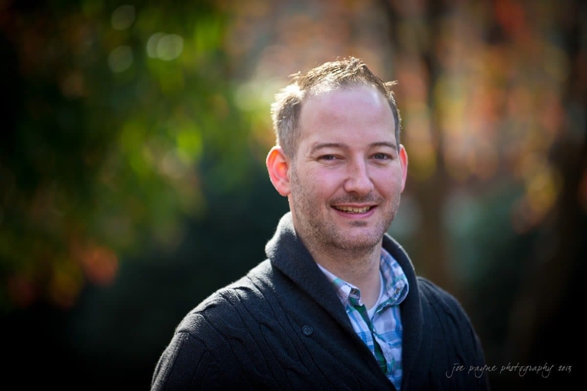 wedding photographers in raleigh nc ~ engagement session for jessica and bo A man in a dark cardigan and plaid shirt smiles at the camera. The background is blurred with greenery and warm tones.