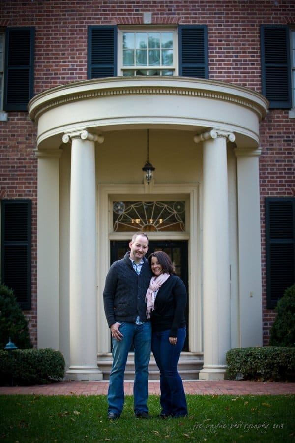 wedding photographers in raleigh nc ~ engagement session for jessica and bo A couple stands in front of a brick building with white columns and a tall door. The man wears a dark sweater and jeans, while the woman is in a gray jacket and jeans, with a pink scarf. Greenery frames the entrance.