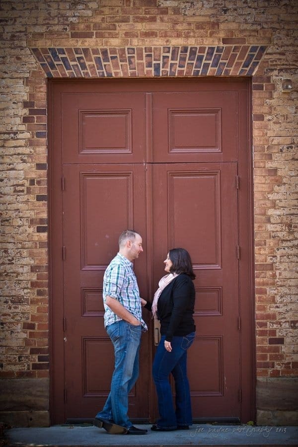 A man and woman stand facing each other, smiling, in front of a large wooden door with a brick wall. The man is wearing a plaid shirt and jeans, and the woman is wearing a black jacket and jeans.