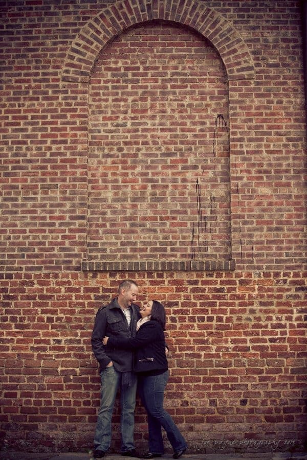 wedding photographers in raleigh nc ~ engagement session for jessica and bo A couple stands close, looking at each other affectionately, in front of a large brick wall. The wall has an arched outline at the top, adding architectural interest to the scene.