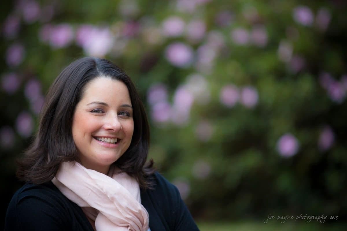 wedding photographers in raleigh nc ~ engagement session for jessica and bo A woman with dark hair smiles while wearing a black top and a light pink scarf. She is outdoors with a blurred background of green foliage and pink flowers.