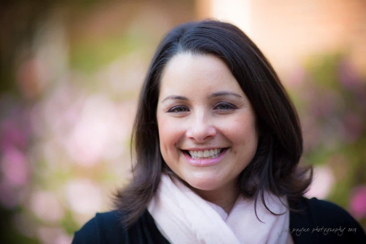 wedding photographers in raleigh nc ~ engagement session for jessica and bo A woman with long brown hair is smiling at the camera. She is wearing a light pink scarf and a dark top. The background is softly blurred with hints of pink and green, suggesting a garden or park setting.