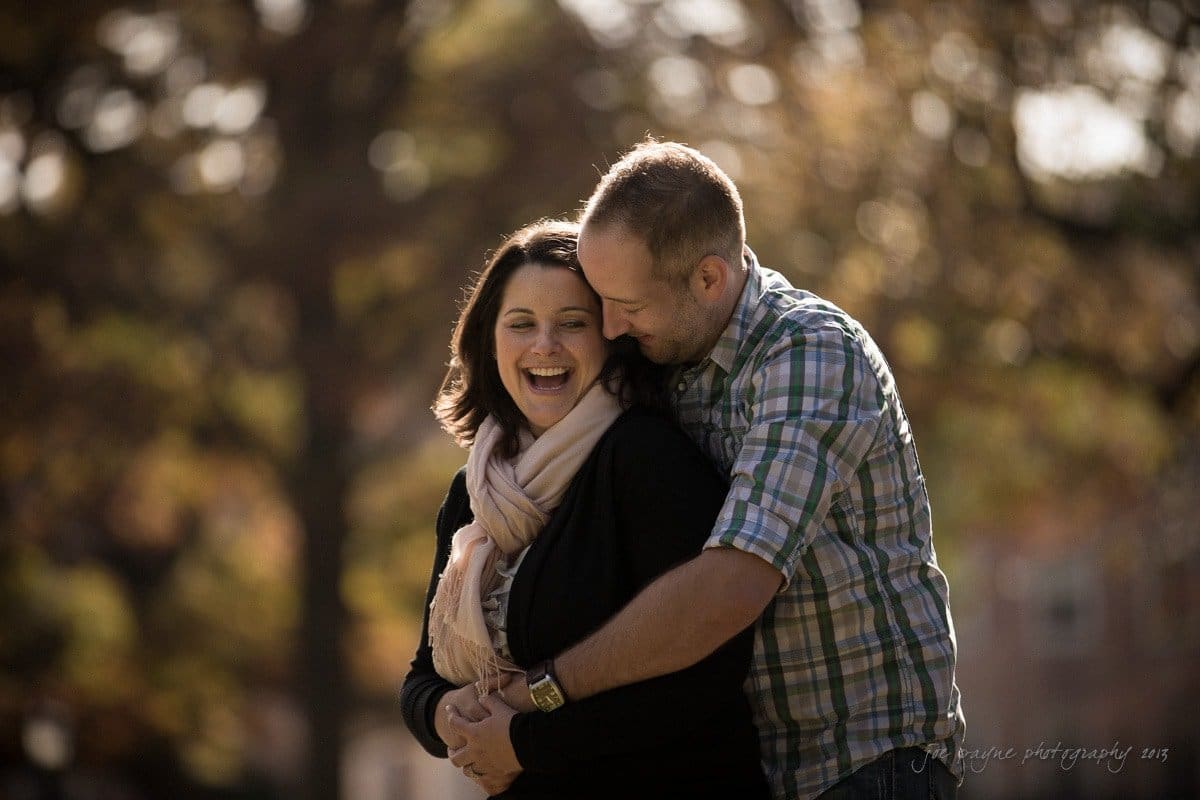 wedding photographers in raleigh nc ~ engagement session for jessica and bo A smiling couple embraces outdoors. The man wears a plaid shirt and leans in close, while the woman, wearing a scarf, laughs. The background is softly blurred with autumnal trees.