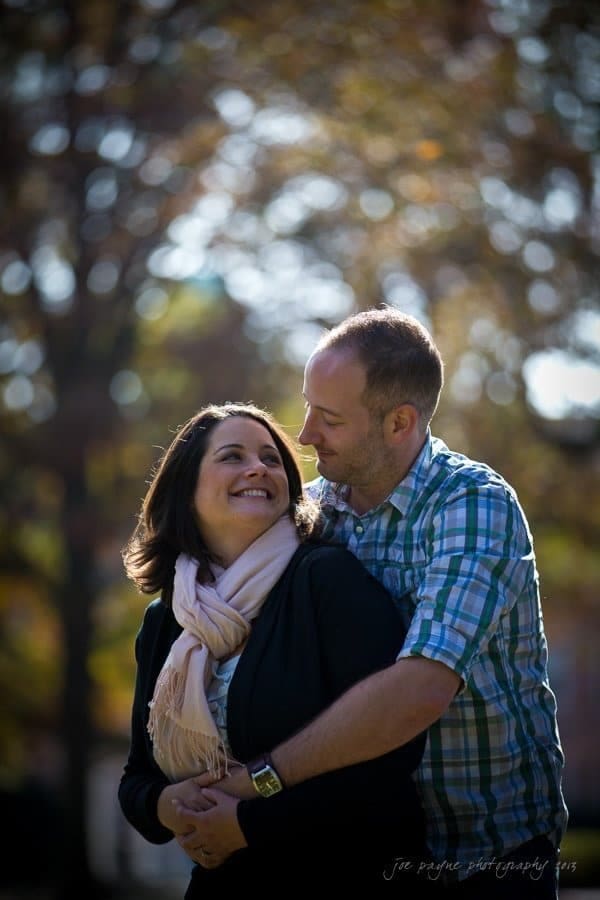 wedding photographers in raleigh nc ~ engagement session for jessica and bo A couple stands closely, with the man embracing the woman from behind. They are both smiling against an outdoor backdrop of trees with sunlight filtering through. The woman wears a scarf, and the man is in a plaid shirt.