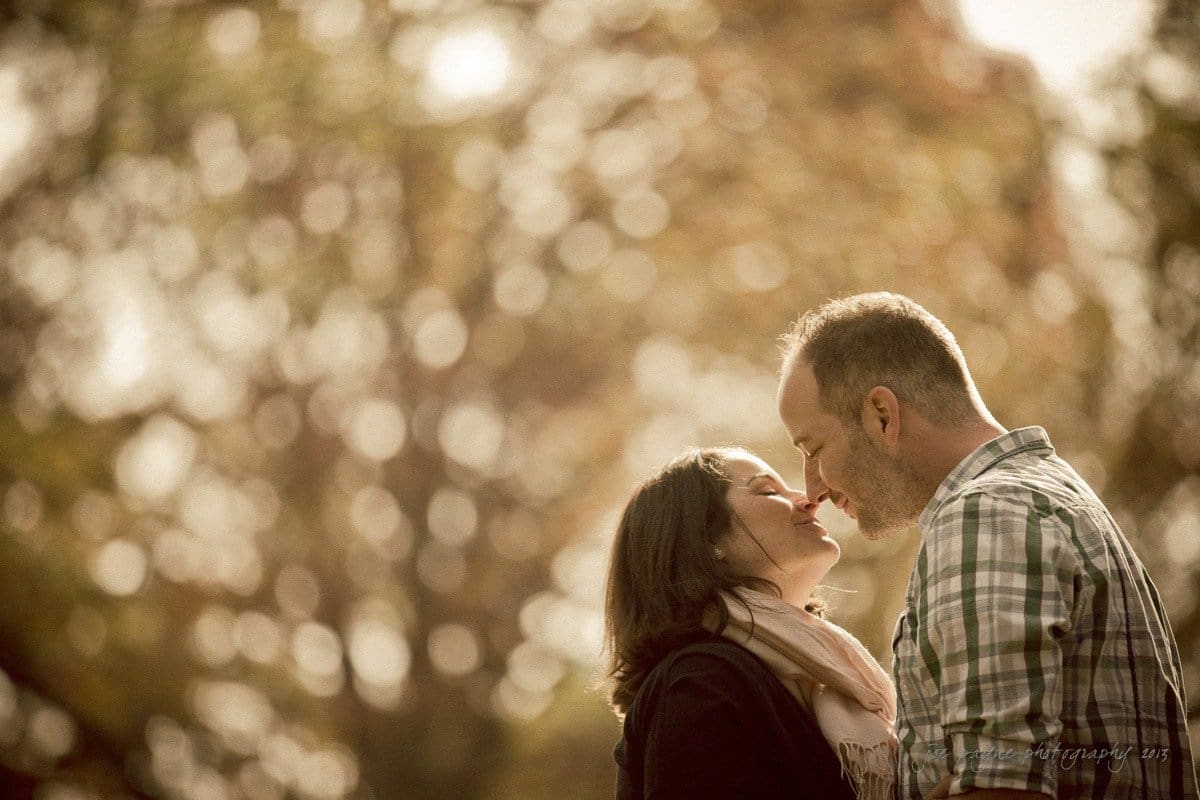 wedding photographers in raleigh nc ~ engagement session for jessica and bo A couple stands closely, touching noses. They are outdoors with an autumnal background of blurred trees and warm light. The woman wears a scarf, and the man is in a plaid shirt. They appear happy and content.