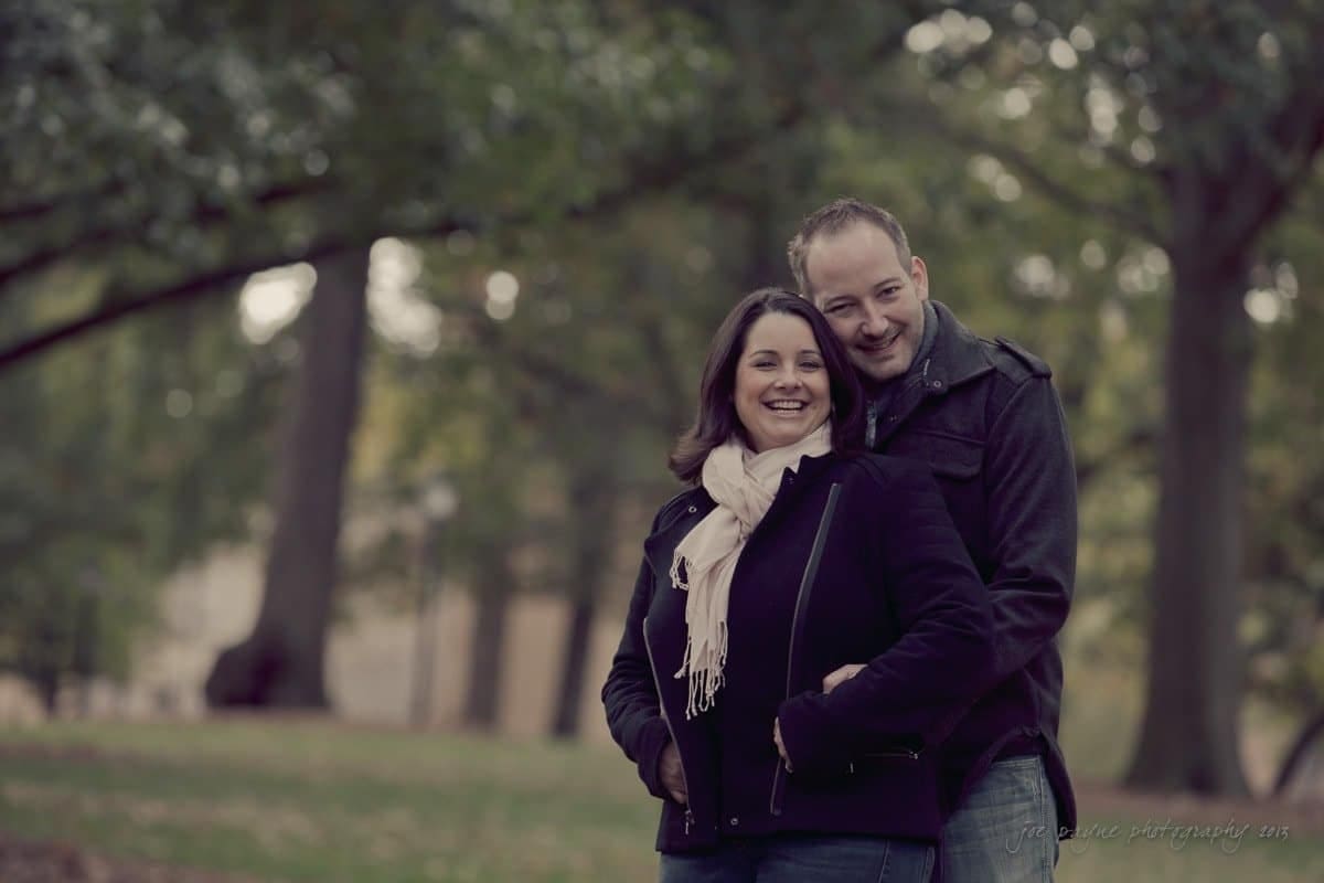 wedding photographers in raleigh nc ~ engagement session for jessica and bo A couple stands in a park on a cloudy day, smiling warmly. The woman is in front, wearing a scarf and dark jacket, while the man stands behind her in a coat, gently embracing her. Trees and a soft background blur frame the scene.