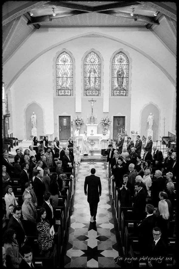 A black and white photograph of a bride and groom standing at an altar in a church. The view is from the back, showing guests seated on either side, with a lone person walking down the aisle towards the altar. Stained glass windows are visible above.