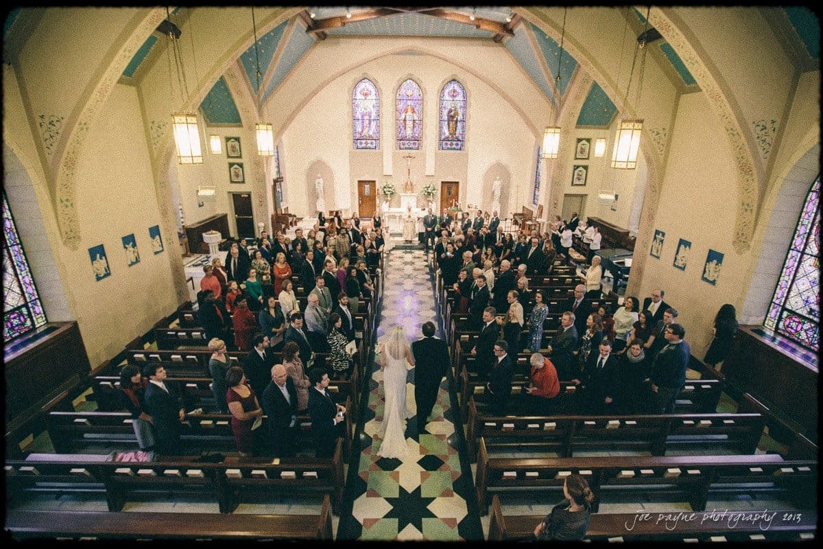 A bride and groom stand at the altar in a church filled with seated guests. The church has vaulted ceilings, stained glass windows, and ornate decor. The view is from above, highlighting the aisle leading to the altar.