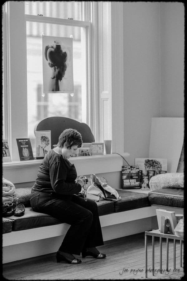 A black and white photo of a woman sitting on a bench in a room. She is looking into a bag on her lap. Behind her is a large window with a portrait and some books and magazines displayed on the windowsill. The room appears relaxed and quiet.