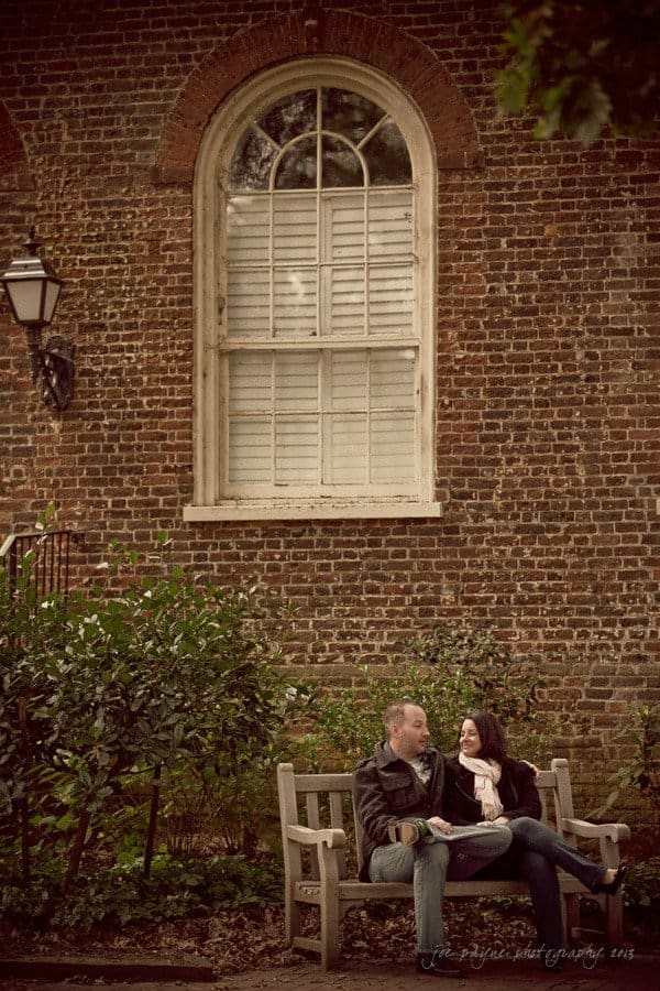 wedding photographers in raleigh nc ~ engagement session for jessica and bo A couple sits on a wooden bench, talking and smiling, in front of a historic brick wall with a large arched window. Greenery surrounds the bench, and a vintage streetlamp hangs on the wall.