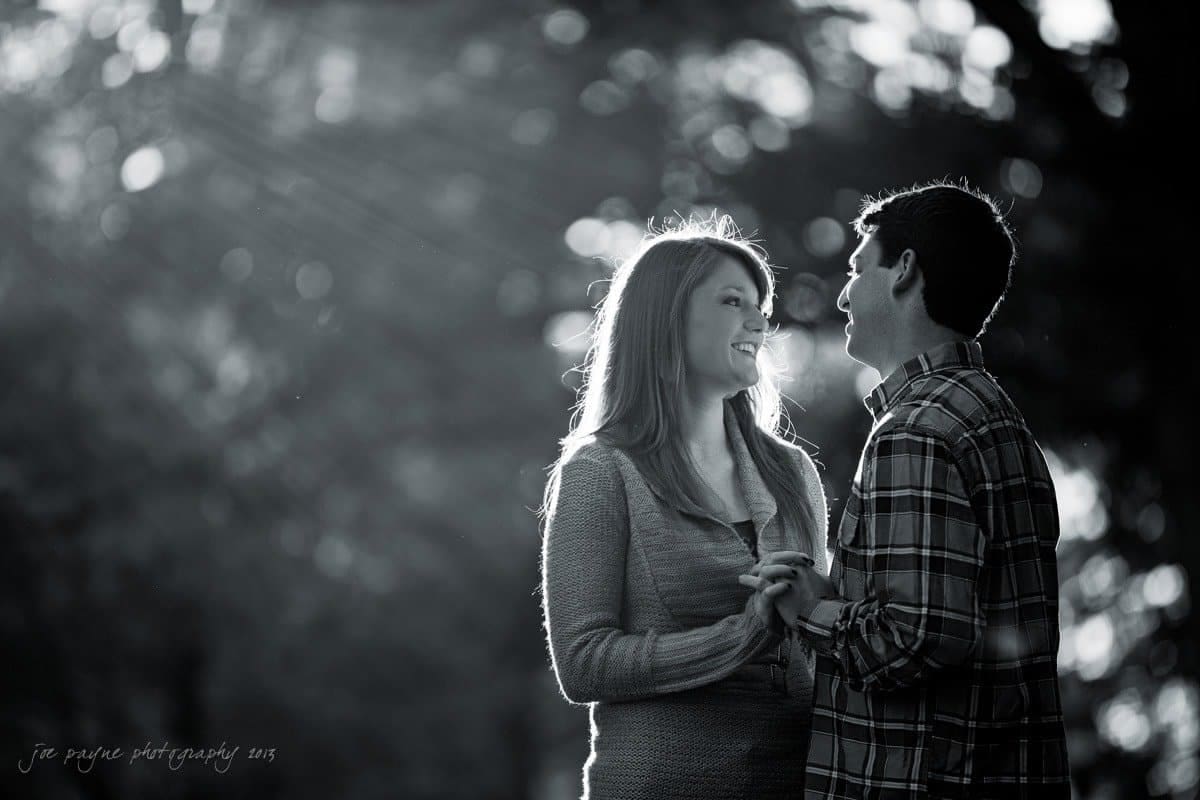 chapel hill engagement session ~ tara and seth A black and white photo of a couple facing each other, smiling and holding hands. Sunlight streams through trees in the background, creating a soft, glowing atmosphere. The woman has long hair and wears a sweater, while the man wears a plaid shirt.