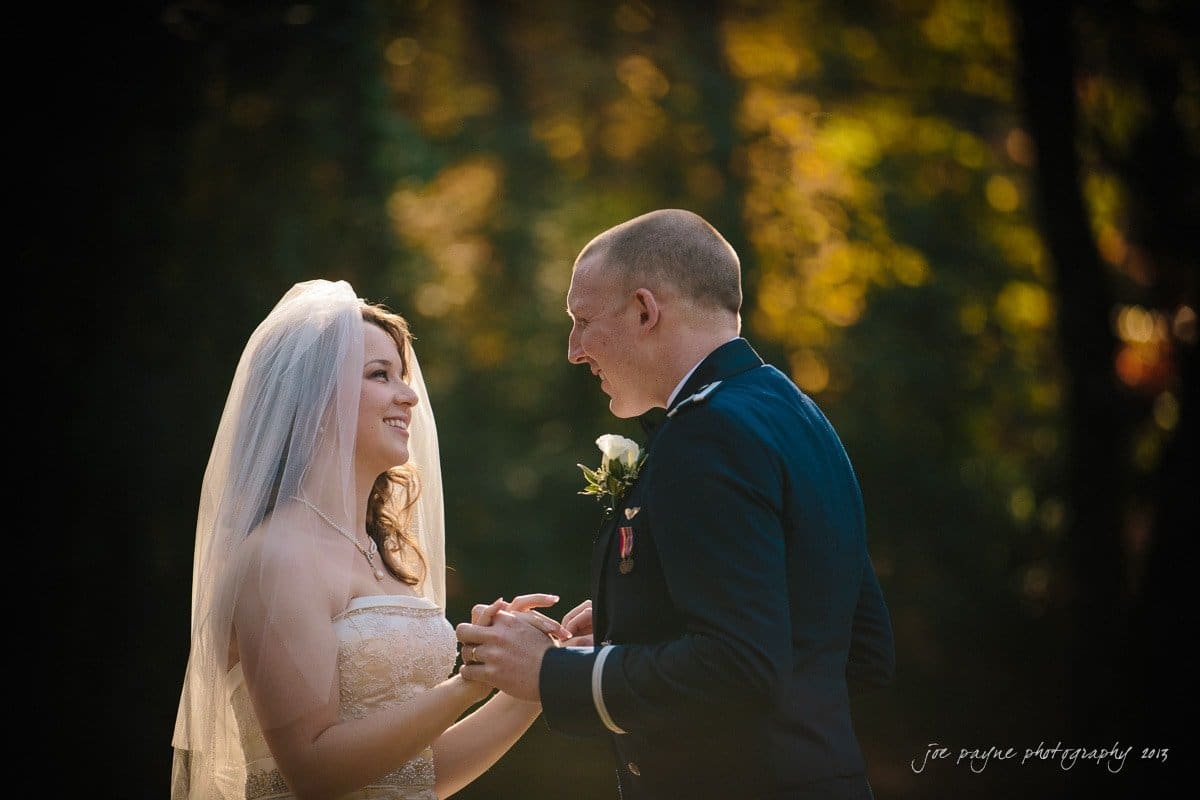 A bride in a white dress and veil smiles at a groom in a dark suit with military-style details. They hold hands and gaze into each others eyes, standing outdoors with a blurred backdrop of trees.