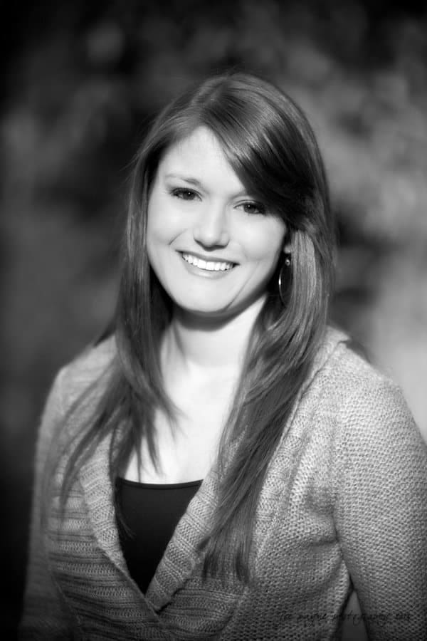 chapel hill engagement session ~ tara and seth A black and white portrait of a smiling woman with long hair, wearing a knit sweater and hoop earrings. The background is softly blurred, emphasizing her face and outfit.
