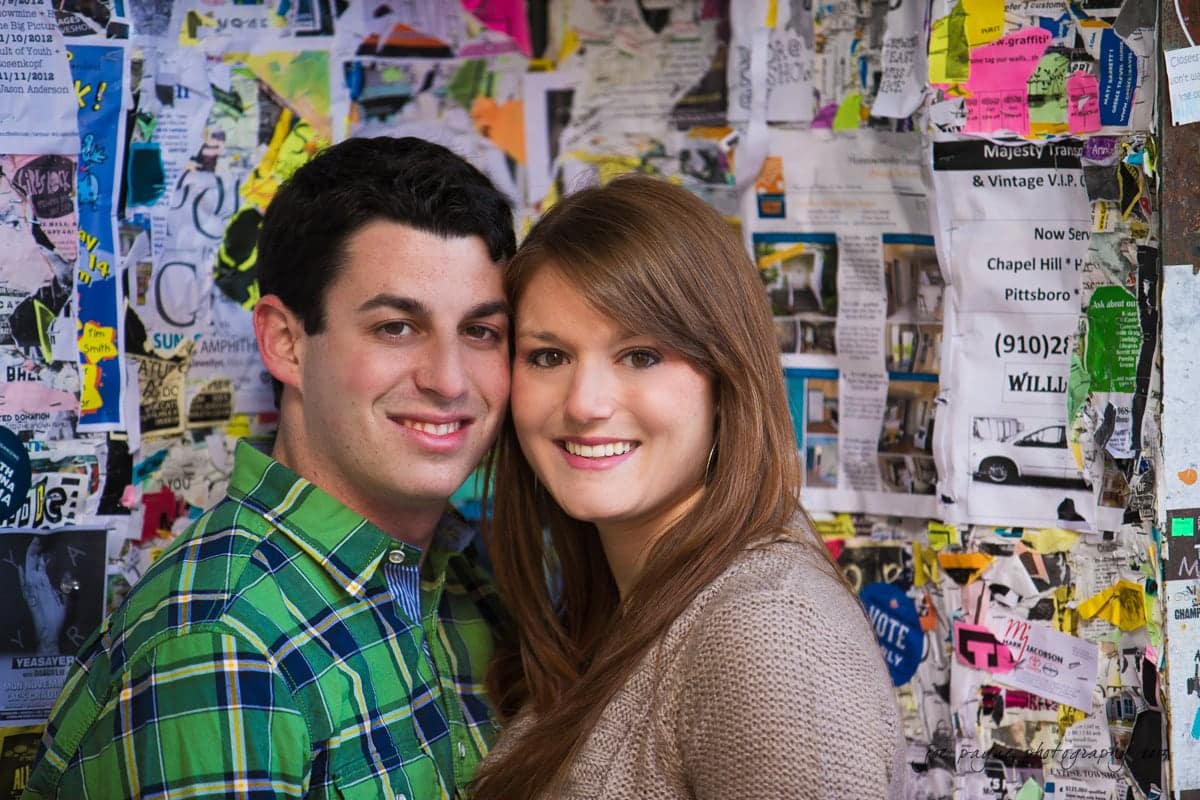 chapel hill engagement session ~ tara and seth A man and woman stand close together, smiling, in front of a colorful bulletin board covered with various flyers and posters. The man wears a green plaid shirt and the woman has long brown hair and wears a beige sweater.