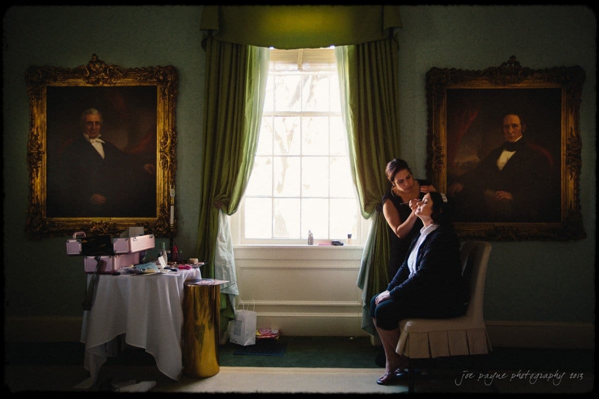 A makeup artist applies makeup to a seated person in a room with elegant decor. Large framed portraits hang on the walls, and a window with green curtains lets in natural light. A small table with various items is on the left.